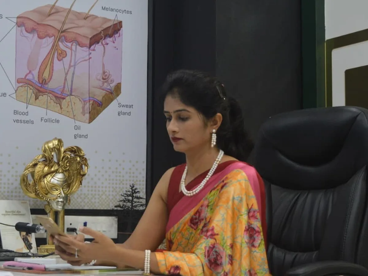 Dr. Daksha Patel during a consultation, with a skin anatomy chart on the wall behind her
